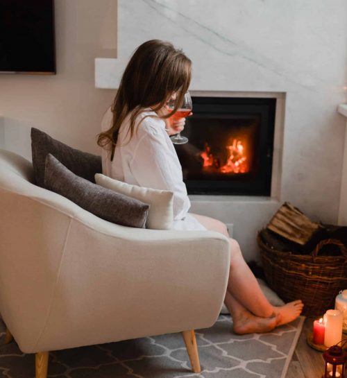 Side view of a young brunette girl is sitting by the fireplace. The beautiful girl is sitting in a cozy room with a glass of red wine